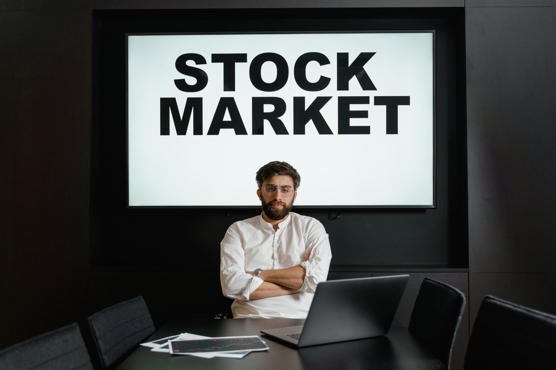Businessman in modern office setting with a stock market display, looking thoughtful and analytical.
