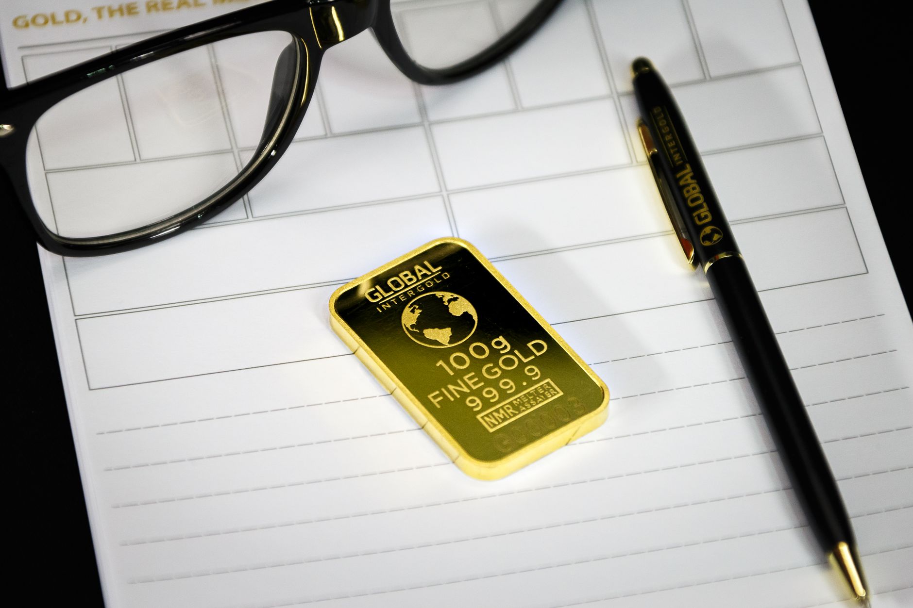 Gold bullion on notepad with eyeglasses and pen, symbolizing wealth and business planning.